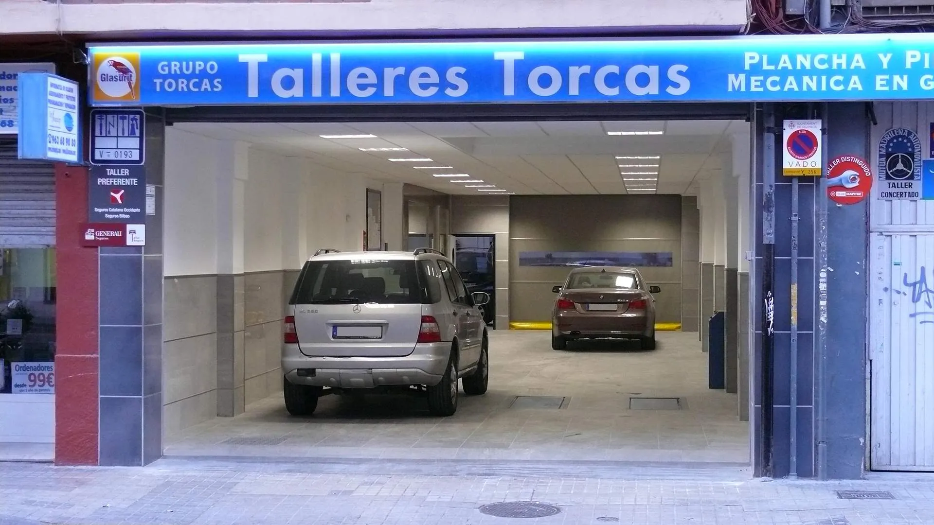 Entrada del taller en Av. Primado Reig 59, con dos coches aparcados dentro de la nave bajo el rótulo de Talleres Torcas.